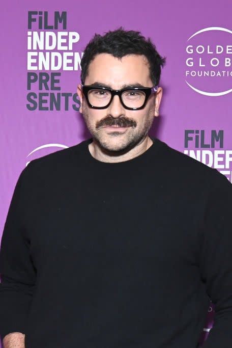 Dan Levy on the red carpet at a Film Independent Presents event, wearing a black sweater and glasses. Golden Globe Foundation logos are displayed in the background