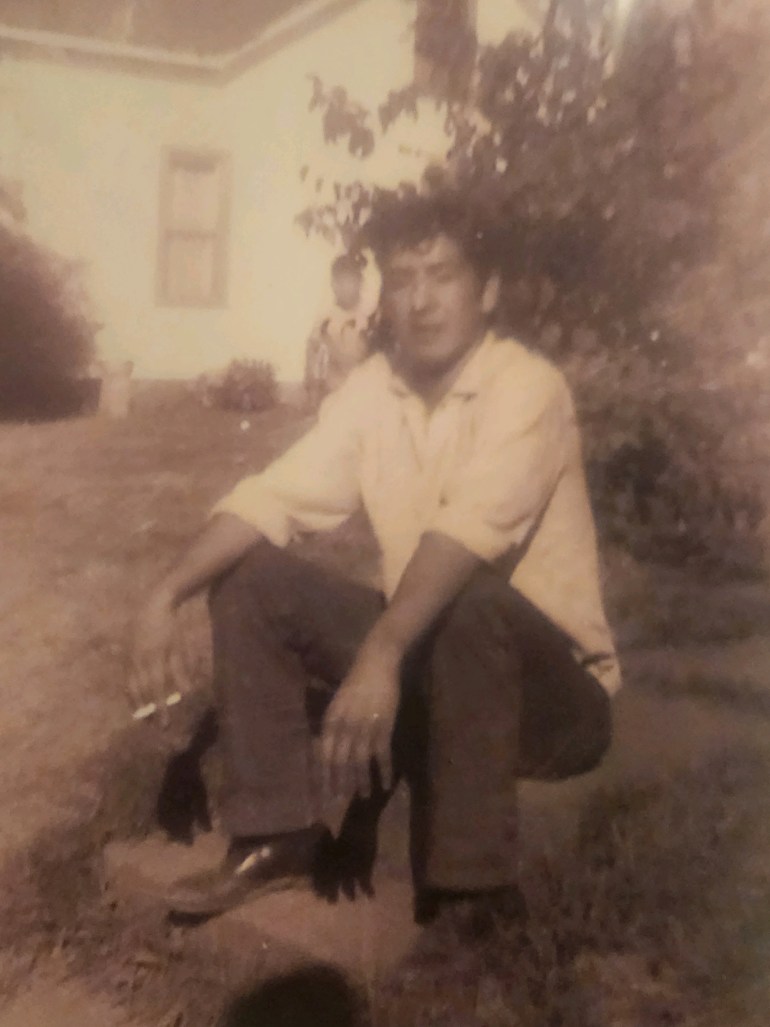 A black-and-white photo of a young Leonard Peltier, sitting on the ground outside in a white collared shirt and black slacks.