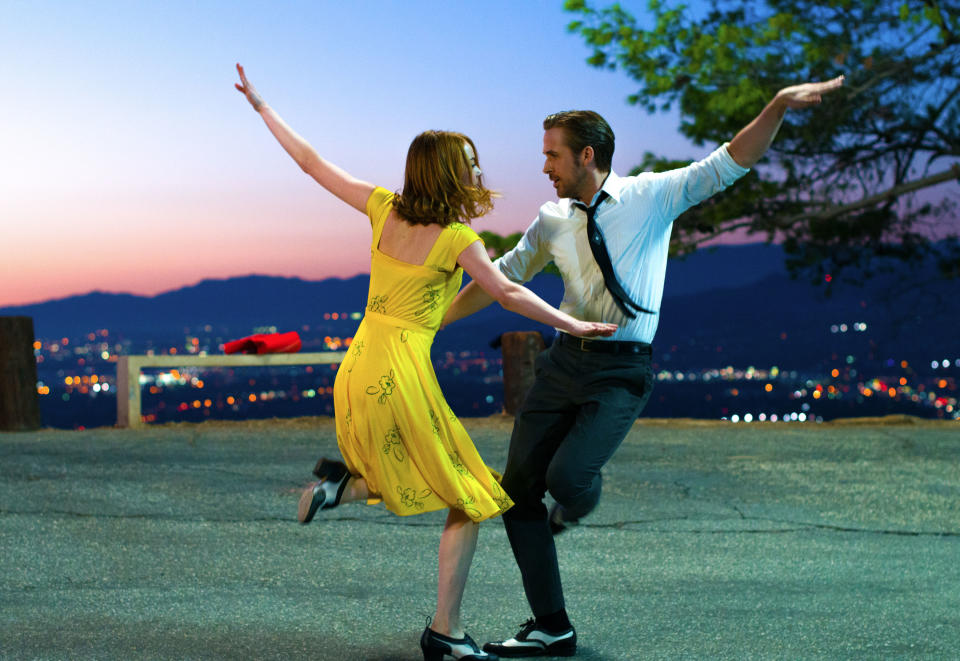 Emma Stone and Ryan Gosling dance joyfully on an overlook in semi-formal attire with a cityscape in the background at dusk