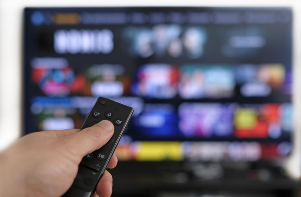 A hand holds a TV remote in front of a large television displaying various streaming service options and shows