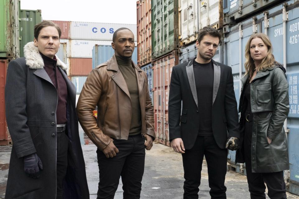 Daniel Brühl, Anthony Mackie, Sebastian Stan, and Emily VanCamp stand in front of shipping containers, dressed in stylish contemporary outfits, looking intense