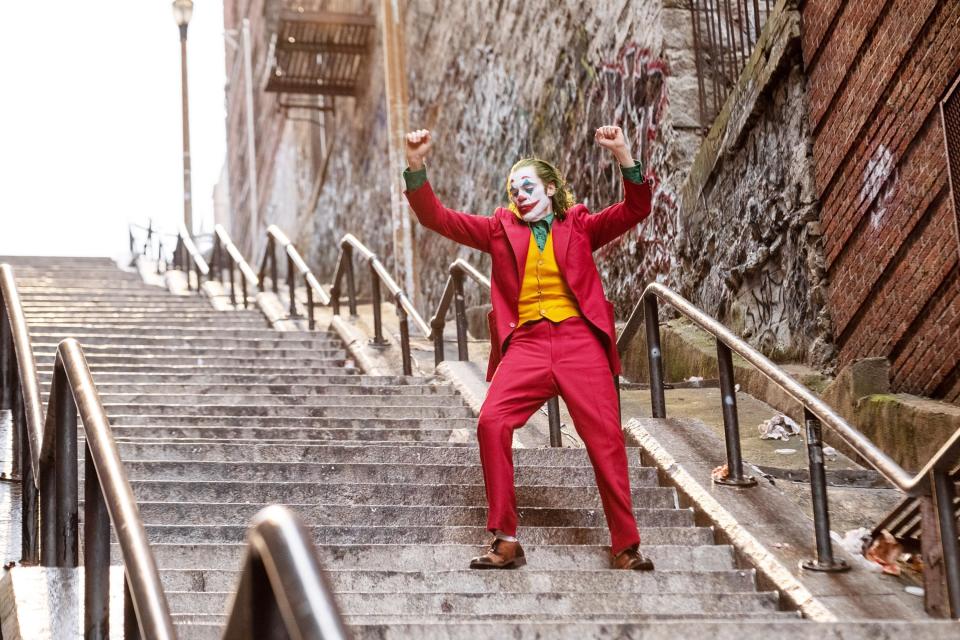 Joaquin Phoenix, dressed as the Joker in a red suit and yellow vest, raises his arms in triumph while dancing on the stairs in a gritty urban setting